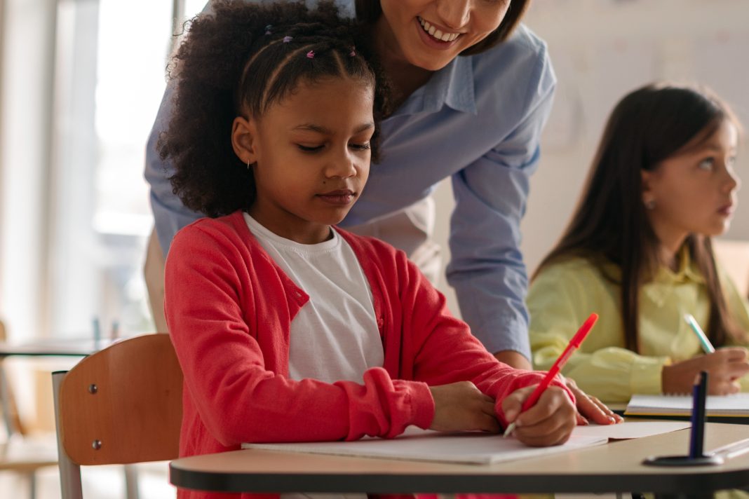 Teacher,Assisting,Young,Student,With,Writing,Exercise,At,Desk,,Supportive éxito